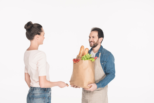 side view of woman giving credit card to shop assistant in apron with paper package full of food isolated on white - Foto, afbeelding