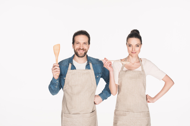 portrait of smiling couple in aprons with wooden kitchen utensils isolated on white - Fotoğraf, Görsel