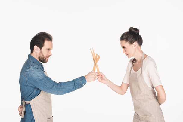 vista lateral do casal em aventais fingindo lutar com utensílios de cozinha de madeira isolados em branco
 - Foto, Imagem
