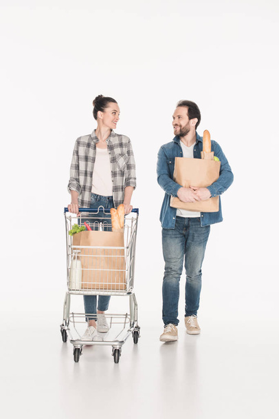 wife and husband with paper packages full of food and shopping trolley looking at each other isolated on white - Photo, Image