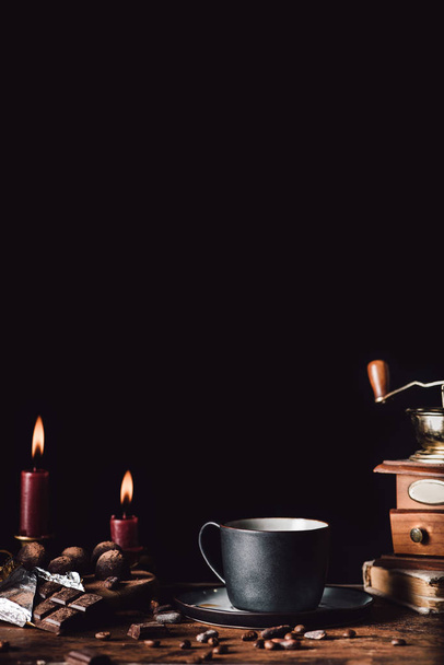 close up image of cup of coffee at wooden table with chocolate, truffles,coffee grains and candles on black background  - Photo, image