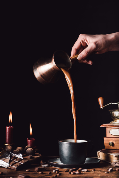 image partielle de la femme versant du café de turc dans une tasse à table avec du chocolat, des truffes, des bougies et des grains de café sur fond noir
 - Photo, image