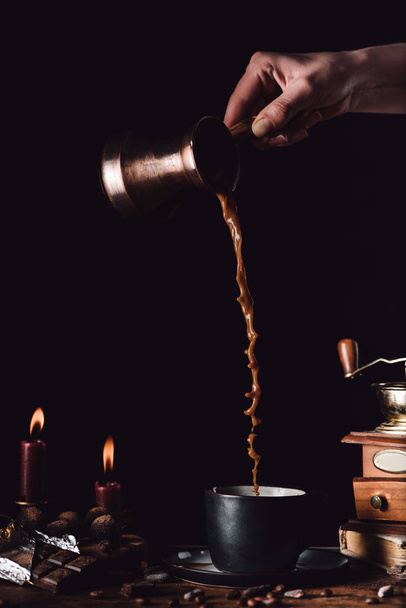 cropped shot of woman pouring coffee from turk into cup at table with chocolate, truffles, candles and coffee grains on black background - Photo, Image