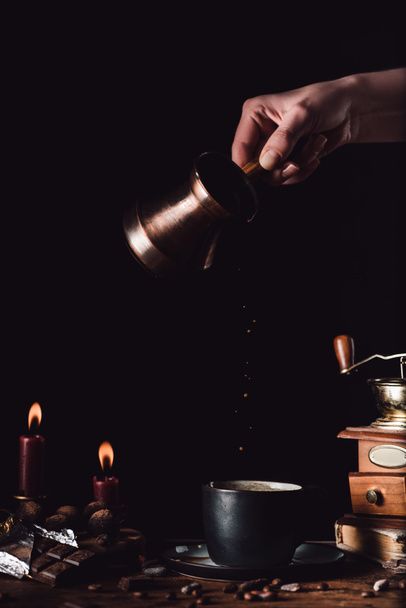 partial view of woman pouring coffee from turk into cup at table with chocolate, truffles, candles and coffee grains on black background - Foto, afbeelding