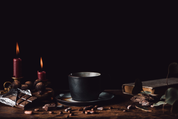 close up shot of cup of coffee at wooden table with chocolate, truffles, coffee grains, candles and book on black background  - Fotografie, Obrázek