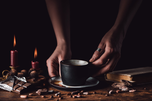 cropped shot of woman holding cup of coffee at wooden table with chocolate, truffles, coffee grains, candles and book on black background  - Fotografie, Obrázek