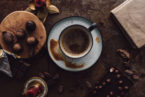 elevated view of cup of coffee, truffles on wooden slice, candles, cocoa beans, books and two chocolate bars on rustic table  - Fotoğraf, Görsel