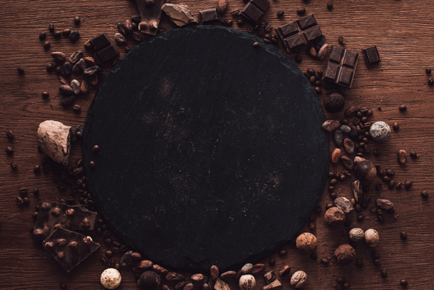 verhoogde weergave van snijplank omringd door verschillende soorten chocoladestukjes, truffels. korrels van koffie en cacao bonen op houten tafel  - Foto, afbeelding