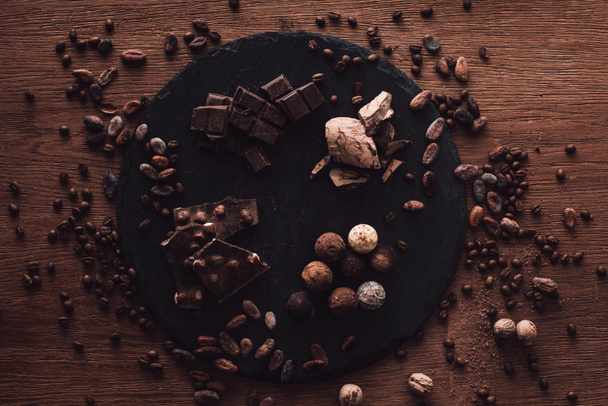elevated view of cutting board with various types of chocolate pieces and truffles surrounded by cocoa beans, coffee grains and nutmegs on wooden table - Photo, Image