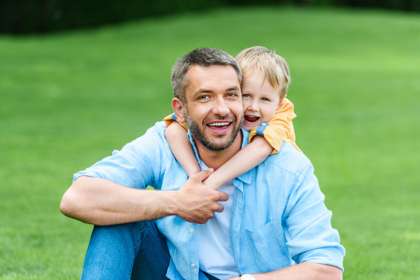 ευτυχισμένος πατέρας και γιος αγκαλιάζει και να χαμογελά στη φωτογραφική μηχανή στο πάρκο - Φωτογραφία, εικόνα