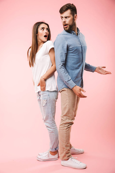 Full length portrait of a confused young couple standing back to back isolated over pink background - Фото, изображение