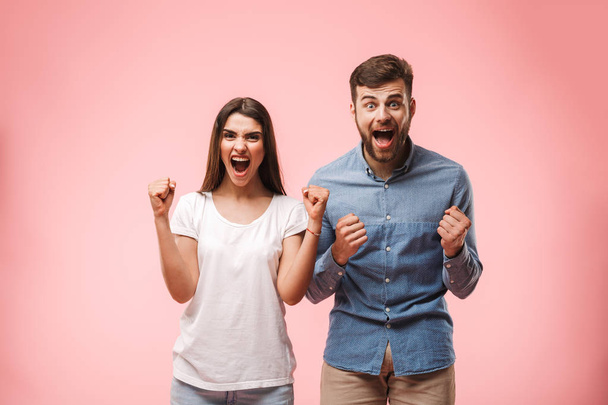 Portrait of an excited young couple screaming and looking at camera isolated over pink background - 写真・画像