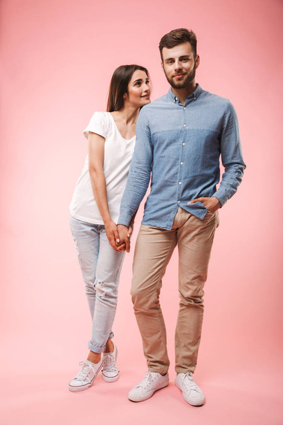 Full length portrait of a smiling young couple standing together holding hands isolated over pink background - Foto, Bild