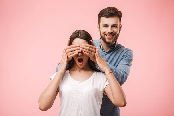 Portrait of a happy young man covering his girlfriends eyes as a surprise isolated over pink background - Fotó, kép