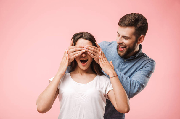 Portrait of a smiling young man covering his girlfriends eyes as a surprise isolated over pink background - Foto, immagini