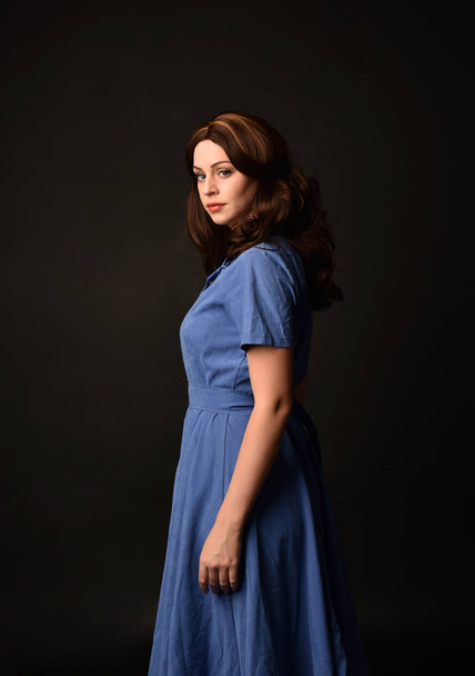 3/4 portret brunetka dama na sobie niebieską sukienkę. pozował z powrotem do aparatu na tle czarny studio. - Zdjęcie, obraz