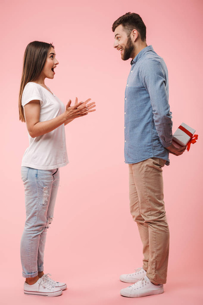 Full length portrait of a happy young man giving his girlfriend a present isolated over pink background - Fotó, kép