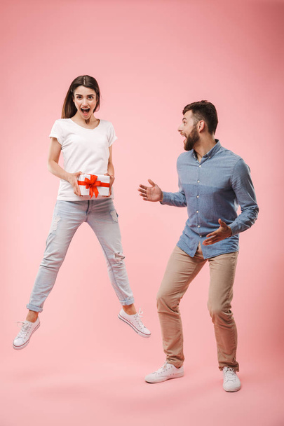 Full length portrait of an excited young couple jumping with a present box isolated over pink background - Φωτογραφία, εικόνα