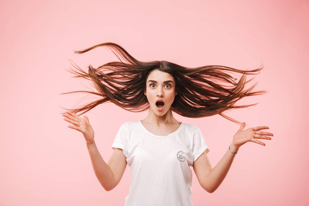 Portrait of a shocked young woman with long hair jumping isolated over pink background - 写真・画像