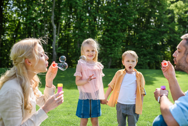 adorable kids holding hands and looking at parents blowing soap bubbles in park - Photo, Image