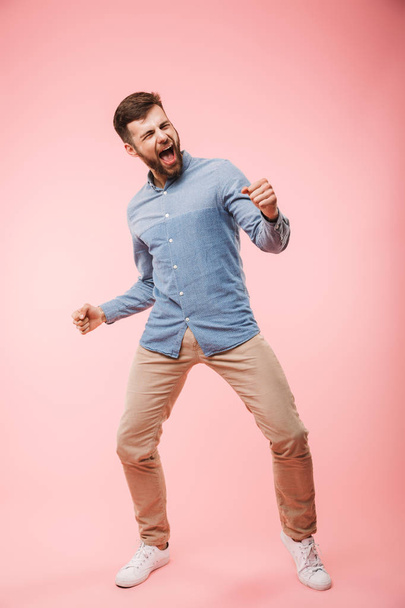 Full length of a cheerful young man standing and celebrating success isolated over pink background - Foto, imagen