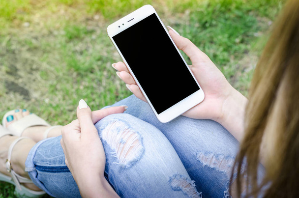 Close Up biały gospodarstwa Smartphone Mobile komórki w kobieta ręce stylowe rozdarty dżinsy długie paznokcie Denim Black Oled wyświetlacz tle natura - Zdjęcie, obraz