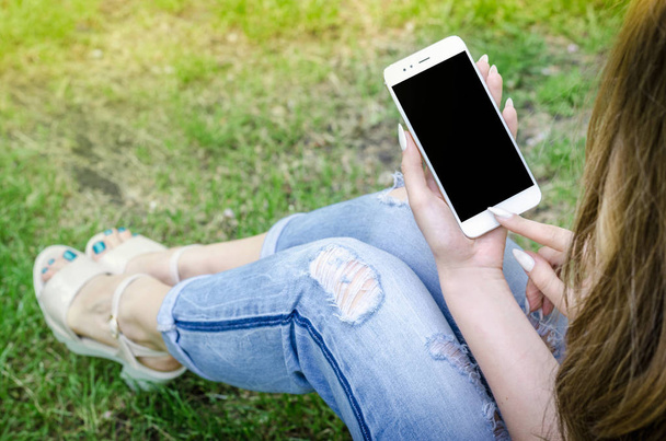 Close Up bílý Smartphone mobilní buňky hospodářství v ženě ruce stylové roztrhané džíny dlouhé Nails Denim Black Oled displej příroda pozadí - Fotografie, Obrázek