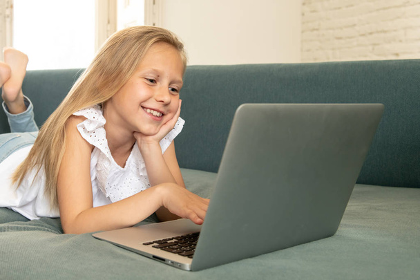 Beautiful cute charming little blonde girl playing and surfing the internet on laptop smiling and lying down on the couch at home. Digital technology, internet usage and life style concept - Fotó, kép