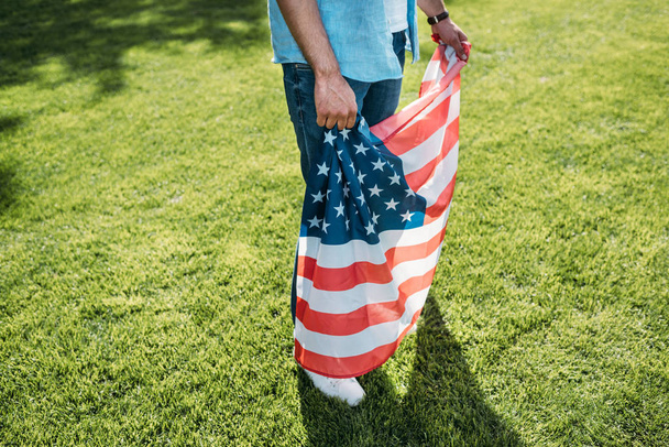 公園の芝生の上に立っている間アメリカの国旗を保持している男のクロップ撮影 - 写真・画像