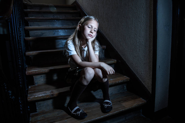 Sad, depressed, unhappy schoolgirl suffering from bullying and abuse feeling lonely and hopeless sitting on stairs with dark light. School isolation, bullying, harassment and abuse concept - Фото, изображение