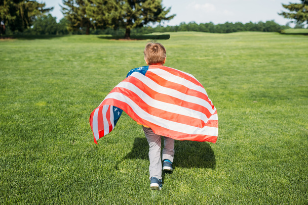 zadní pohled na malého chlapce s americkou vlajkou, běžící na trávě  - Fotografie, Obrázek
