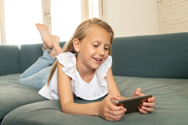 Beautiful cute charming little blonde girl playing and having fun on her smart phone smiling and lying down on the couch at home. Digital technology, mobile usage and life style concept - Zdjęcie, obraz