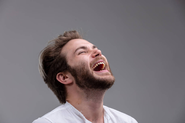 Διασκέδαση νεαρός άνδρας με μια αίσθηση του χιούμορ που απολαμβάνουν ένα εγκάρδιο γέλιο με το κεφάλι ριγμένο πίσω και το στόμα ανοικτό απομονώνονται σε γκρι - Φωτογραφία, εικόνα