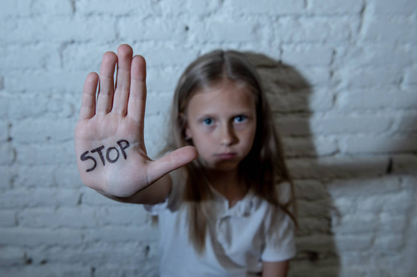 Young cute scared little schoolgirl showing the text stop written in her hand. Children abused and bullied in school concept and children rights. Dramatic light - 写真・画像