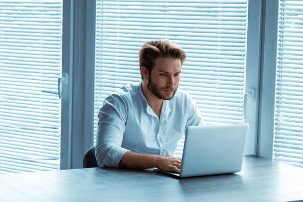 Привлекательный человек, сидящий за ноутбуком за рабочим столом и печатающий информацию перед большими окнами с копировальным местом
 - Фото, изображение