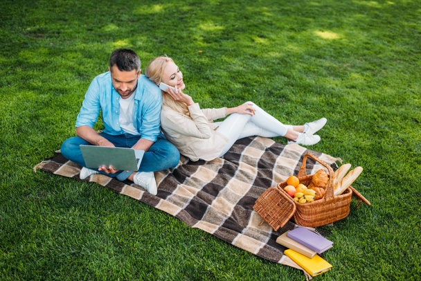 high angle view of happy couple using smartphone and laptop while sitting on plaid at picnic - Фото, зображення