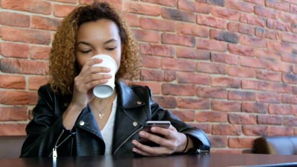 Una hermosa joven negra en una chaqueta de cuero con un vaso blanco de café y teléfono móvil se sienta en una mesa en el fondo de una pared de ladrillo. Enfoque y nitidez en la mano con un smartphone
. - Metraje, vídeo