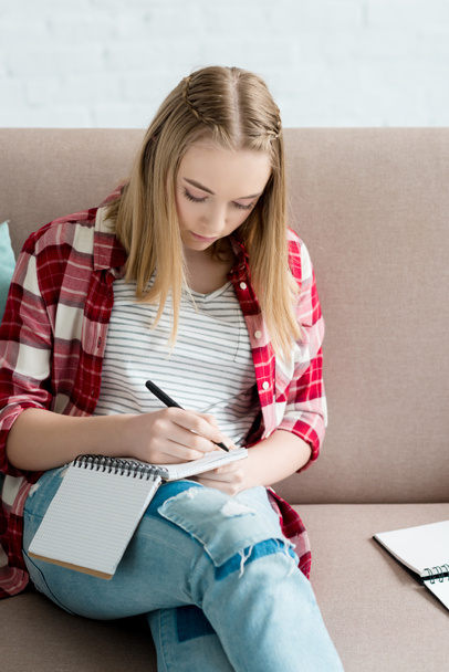 teen student girl writing in notebook while sitting on sofa and doing homework - Foto, imagen