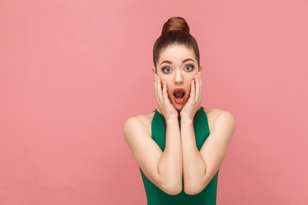 shocked woman wearing green dress with opened mouth touching hands cheeks on pink background - Photo, Image