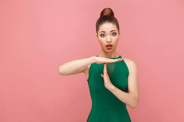 beautiful woman with collected hair in green dress showing time out sign at camera on pink background,  Expression emotion and feelings concept  - Zdjęcie, obraz