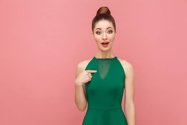 Optimistic successful beautiful woman with collected hair in green dress pointing finger himself on pink background, Expression emotion and feelings concept  - Foto, Bild
