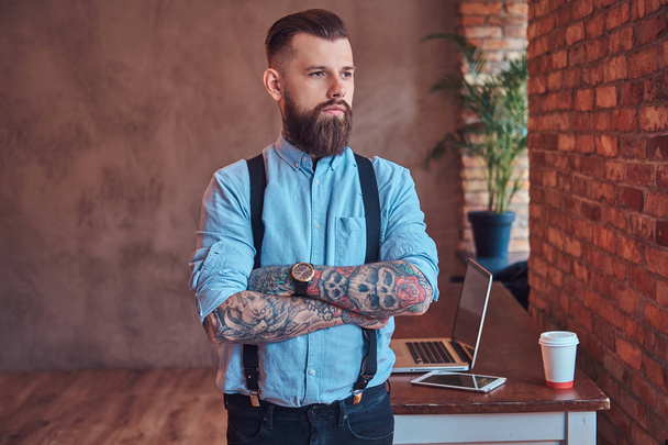 hipster tatoué à l'ancienne dans une chemise et des bretelles, debout près d'un bureau avec un ordinateur portable, regardant par la fenêtre dans un bureau avec un intérieur loft
. - Photo, image
