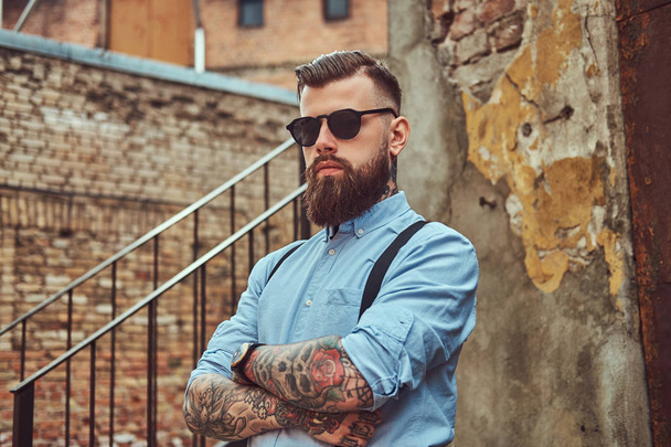 Κομψό όμορφος ντεμοντέ τατουάζ hipster guy σε ένα πουκάμισο με ζαρτιέρες, στέκεται κοντά σε ένα παλιό κτίριο σε εξωτερικούς χώρους. - Φωτογραφία, εικόνα