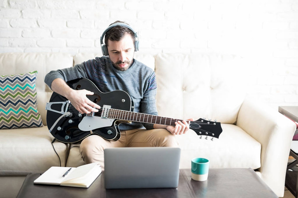 Δημιουργική Λατινική νεαρός με ακουστικά που παίζει μουσική στην κιθάρα και κοιτάζοντας το laptop - Φωτογραφία, εικόνα