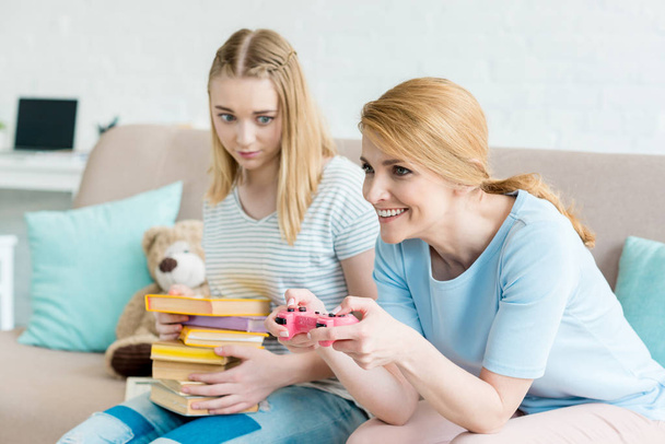 mother playing console game while her confused daughter sitting with stack of books on couch - Photo, Image
