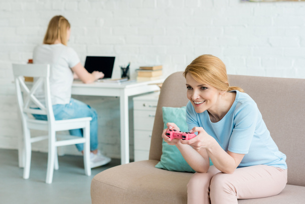 mother playing console game while her daughter working with laptop blurred on background - 写真・画像