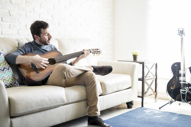 Retrato de belo jovem hispânico sentado no sofá e tocando guitarra
 - Foto, Imagem
