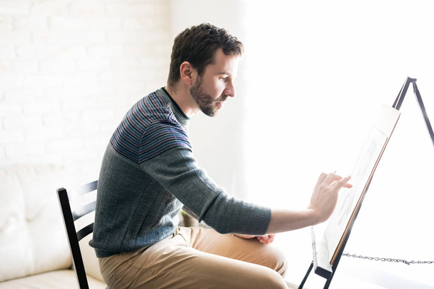 Το προφίλ άποψη του όμορφος Ισπανόφωνος αρσενικός καλλιτέχνης κάνει μια ζωγραφική σε στούντιο στο σπίτι - Φωτογραφία, εικόνα