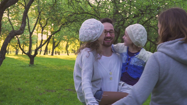 Familia feliz abrazándose en la hierba. Los padres y sus dos hijas se abrazan en el césped del parque. Huerto con flores
. - Imágenes, Vídeo