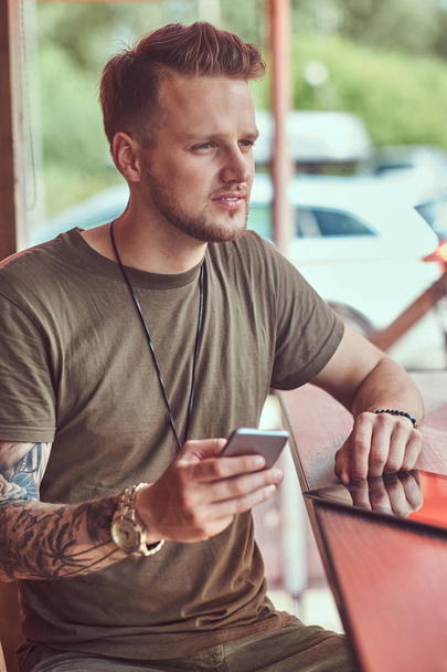 Красивый стильный хипстер сидит за столом в придорожном кафе, держит смартфон
. - Фото, изображение